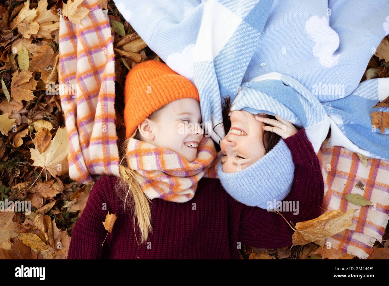 Eine lächelnde, süße Frau und ein kleines Mädchen in warmen Kleidern, die sich anschauen, während sie im Wald oder Park auf Laub liegen Stockfoto