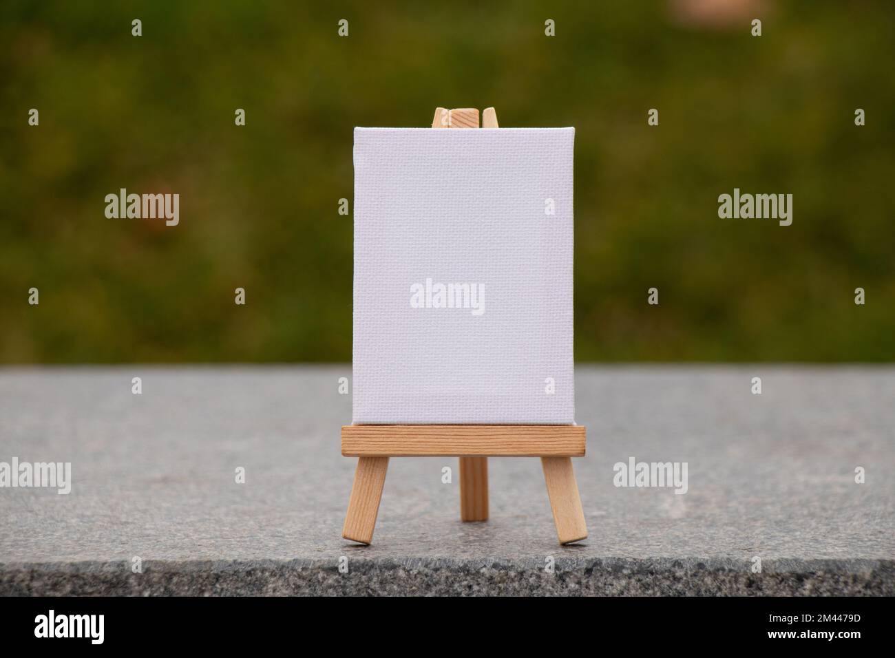 Eine hölzerne Staffelei mit weißem Gemälde steht auf einem grauen Stein auf grünem Hintergrund, Platz für Text Stockfoto