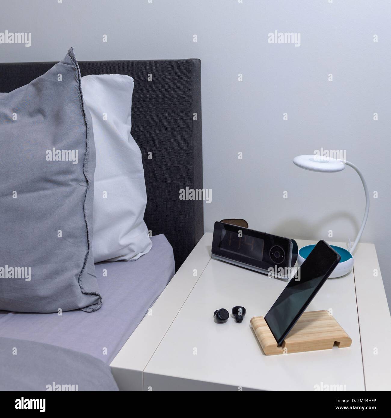 Smartphone auf dem Nachttisch mit Lampe, Headset, Uhr und Bett als Hintergrund. Quadratisches Format Stockfoto