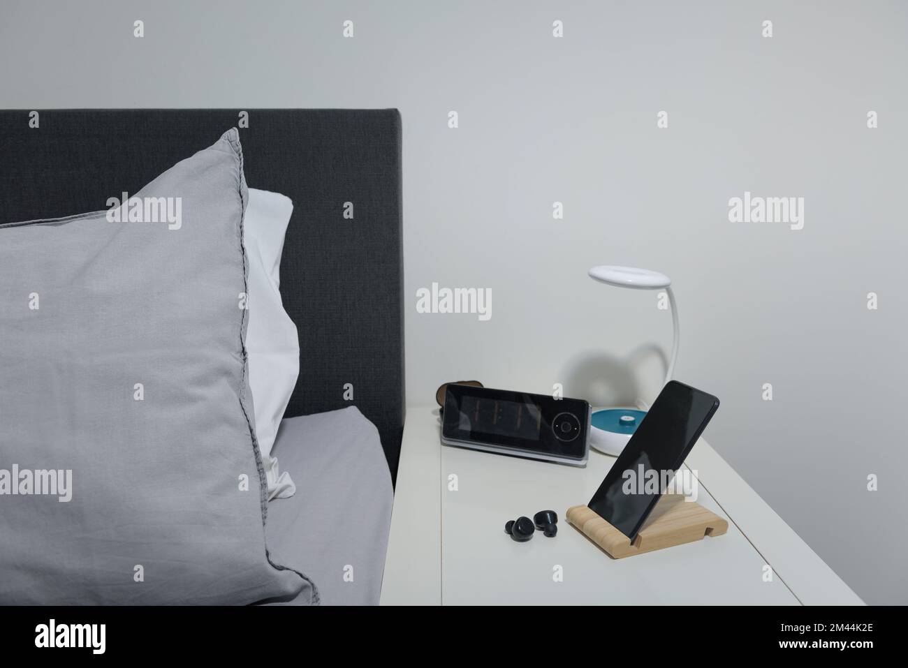 Für das Schlafzimmer. Smartphone auf dem Nachttisch mit Lampe, Headset, Uhr und Bett. Stockfoto