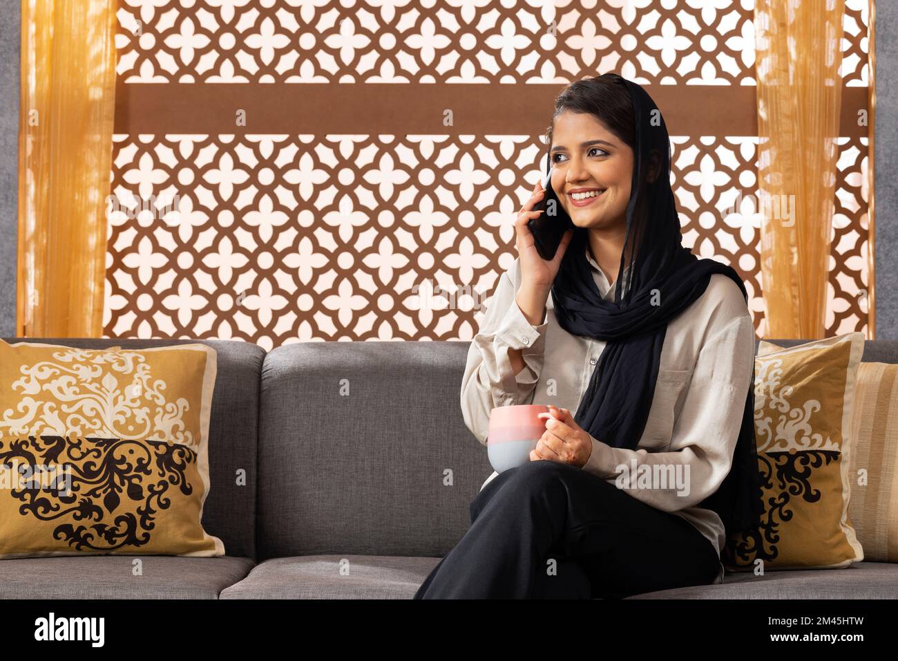 Muslimische Frau, die zu Hause auf dem Sofa sitzt und mit dem Handy telefoniert Stockfoto