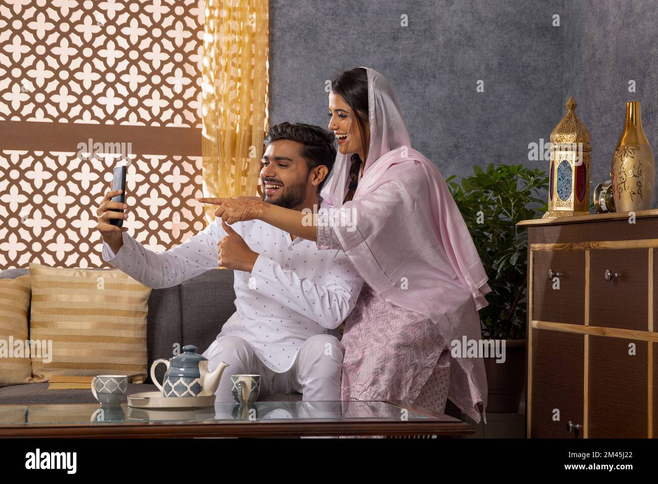 Ein muslimisches Paar, das sich im Wohnzimmer über ein Mobiltelefon per Videoanruf unterhält Stockfoto