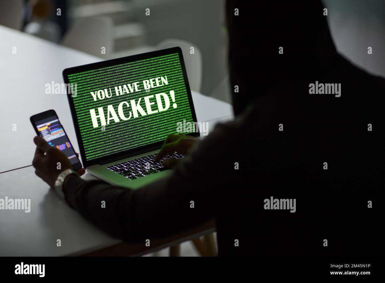 Cyberkriminalität ist eine echte Bedrohung. Ein nicht wiedererkennbarer Hacker, der einen Laptop und ein Smartphone verwendet. Stockfoto