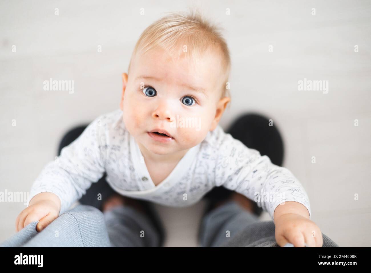 Blick von oben auf den fröhlichen kleinen Jungen, der die ersten Schritte unternimmt und zu Hause an Vaters Trainingshose festhält. Süßer kleiner Junge, der Laufen lernt. Stockfoto