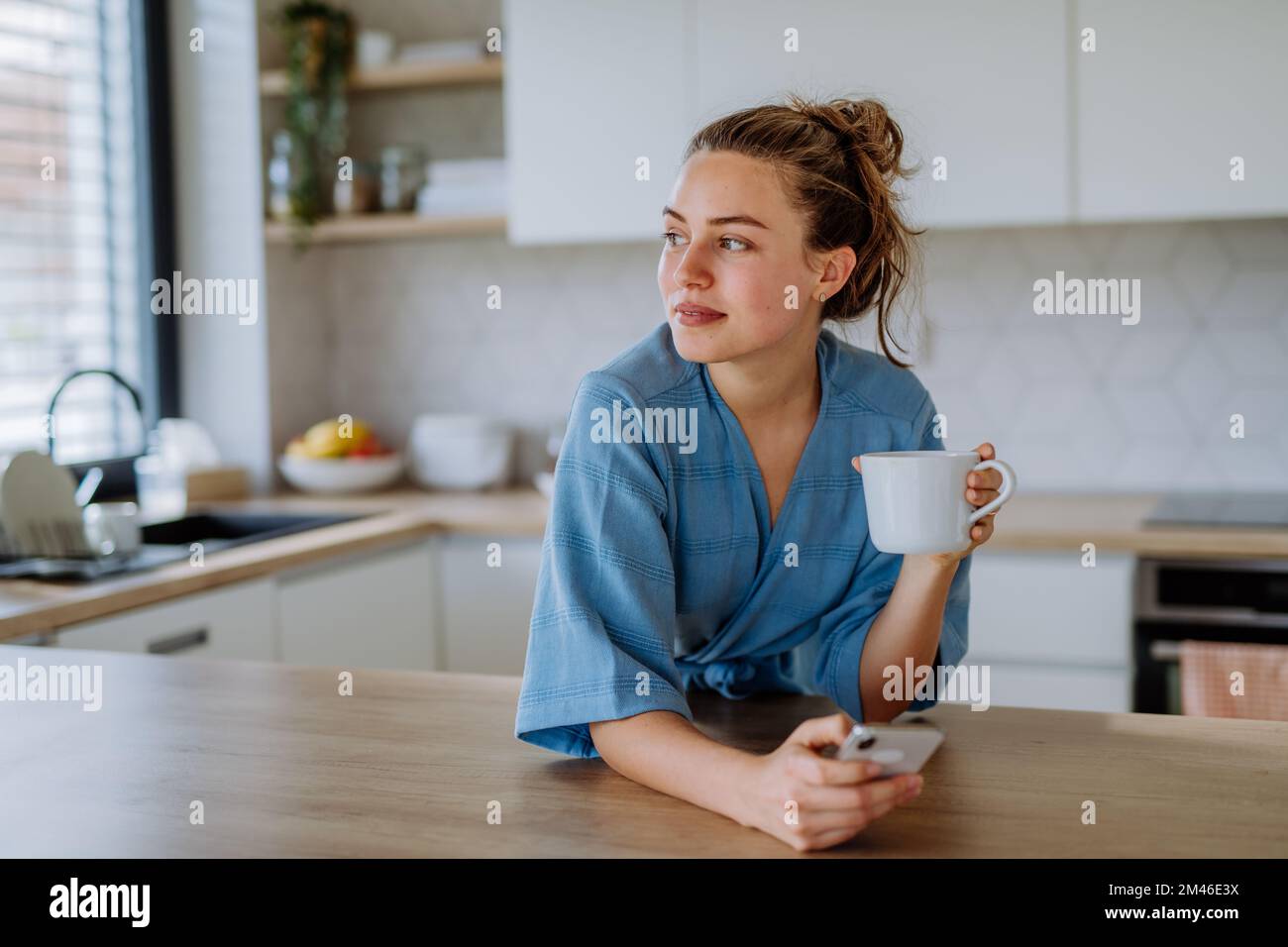 Junge Frau mit Smartphone, die morgens in ihrer Küche eine Tasse Kaffee genießt. Stockfoto