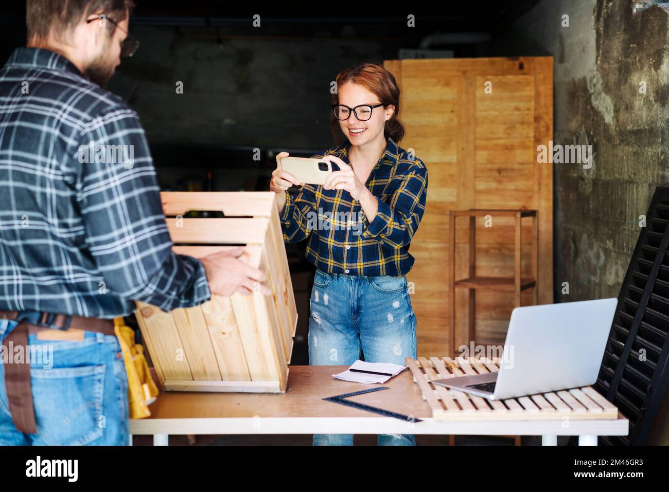Junge Geschäftsfrau, die mit ihrem Smartphone Fotos von Holzprodukten macht, während sie in ihrer eigenen Werkstatt arbeitet. Stockfoto