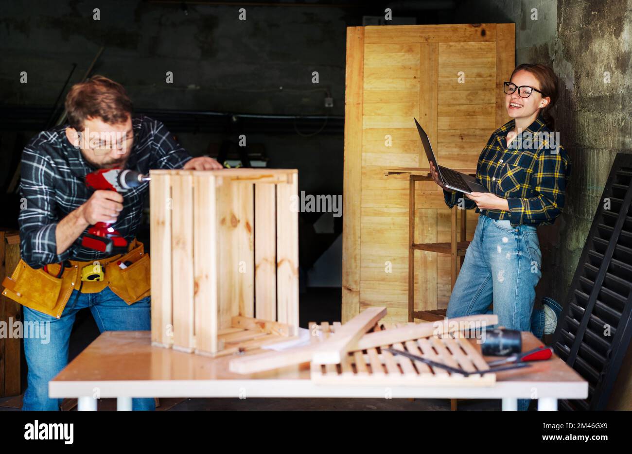 Ein junges Paar, das in seinem kleinen Geschäft zusammenarbeitet, Tischlermann, der eine Frau aus Holzkisten mit einem Laptop macht. Stockfoto