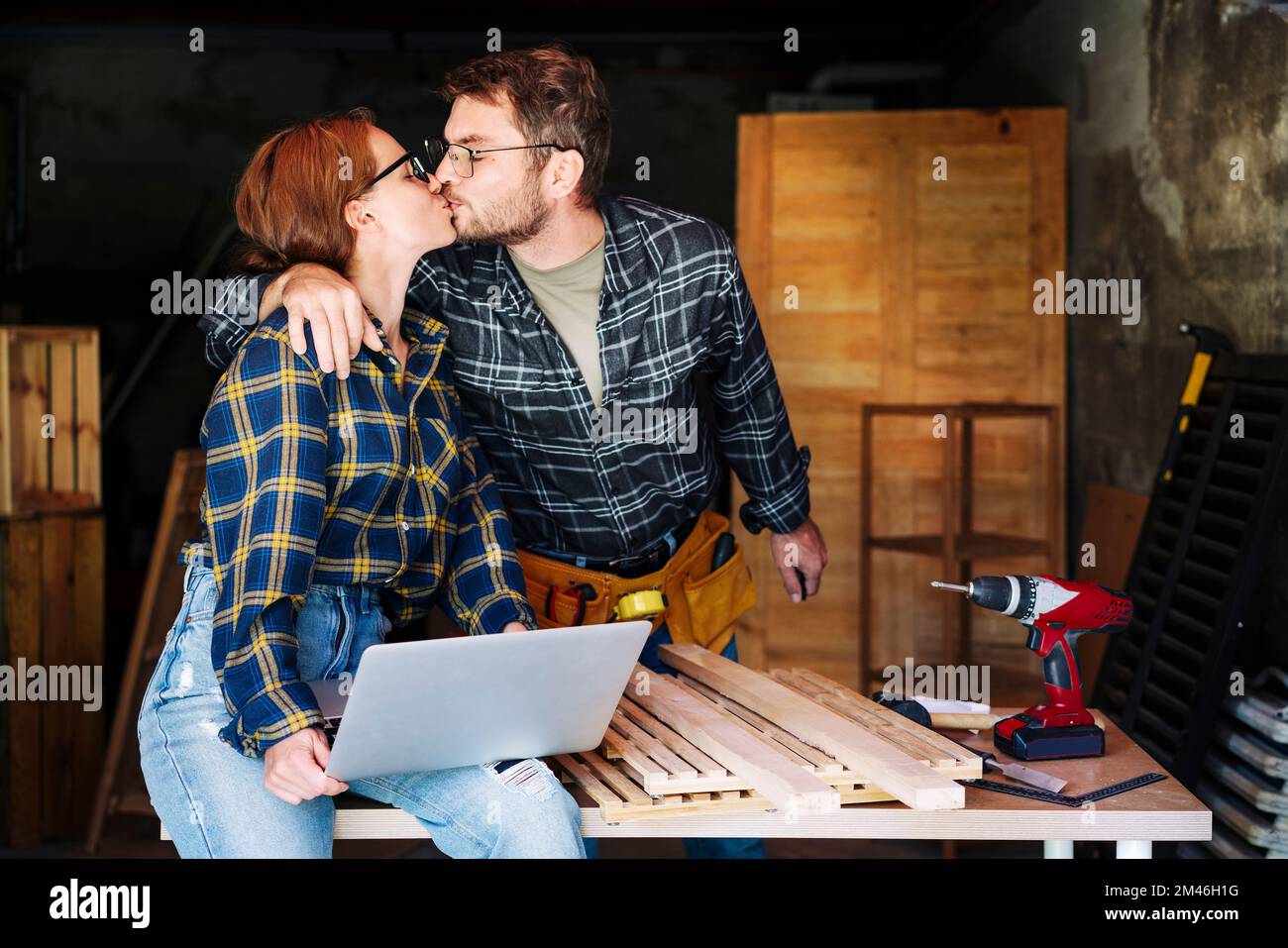 Junge Erwachsene Paare, die sich küssen und den Erfolg ihres kleinen Unternehmens feiern, während sie in ihrer Zimmereiwerkstatt arbeiten. Stockfoto