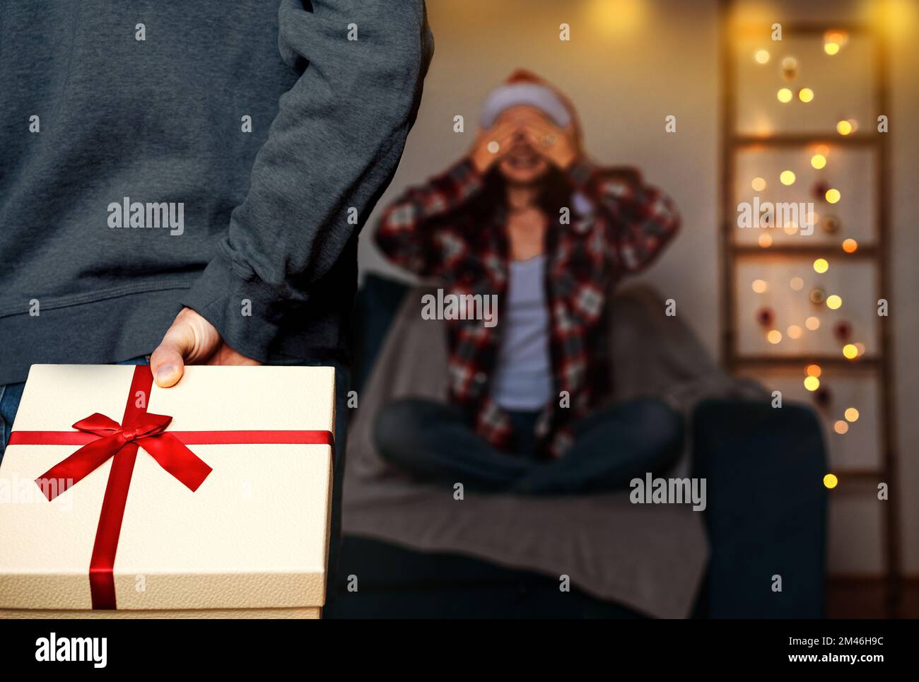 Weihnachtsgeschenke in der Hand hinter dem Rücken unscharfe Frau mit geschlossenen Augen sitzt auf dem Sofa und wartet auf Weihnachtsgeschenke. Stockfoto