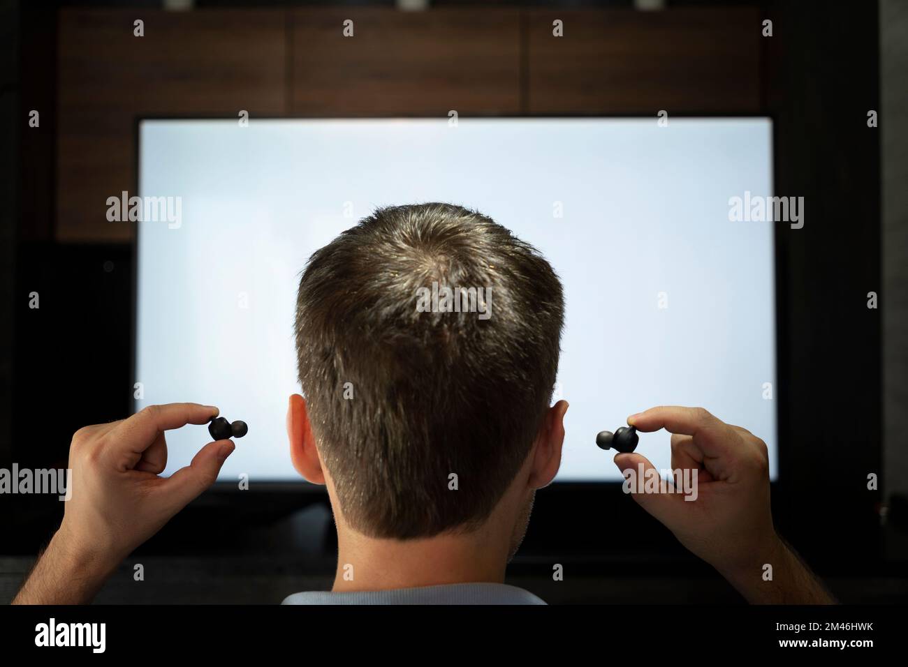 Ein Mann steckt Kopfhörer in sein Ohr vor den Hintergrund eines Fernsehbildschirms. Nachts Fernsehen mit Kopfhörern. Stockfoto