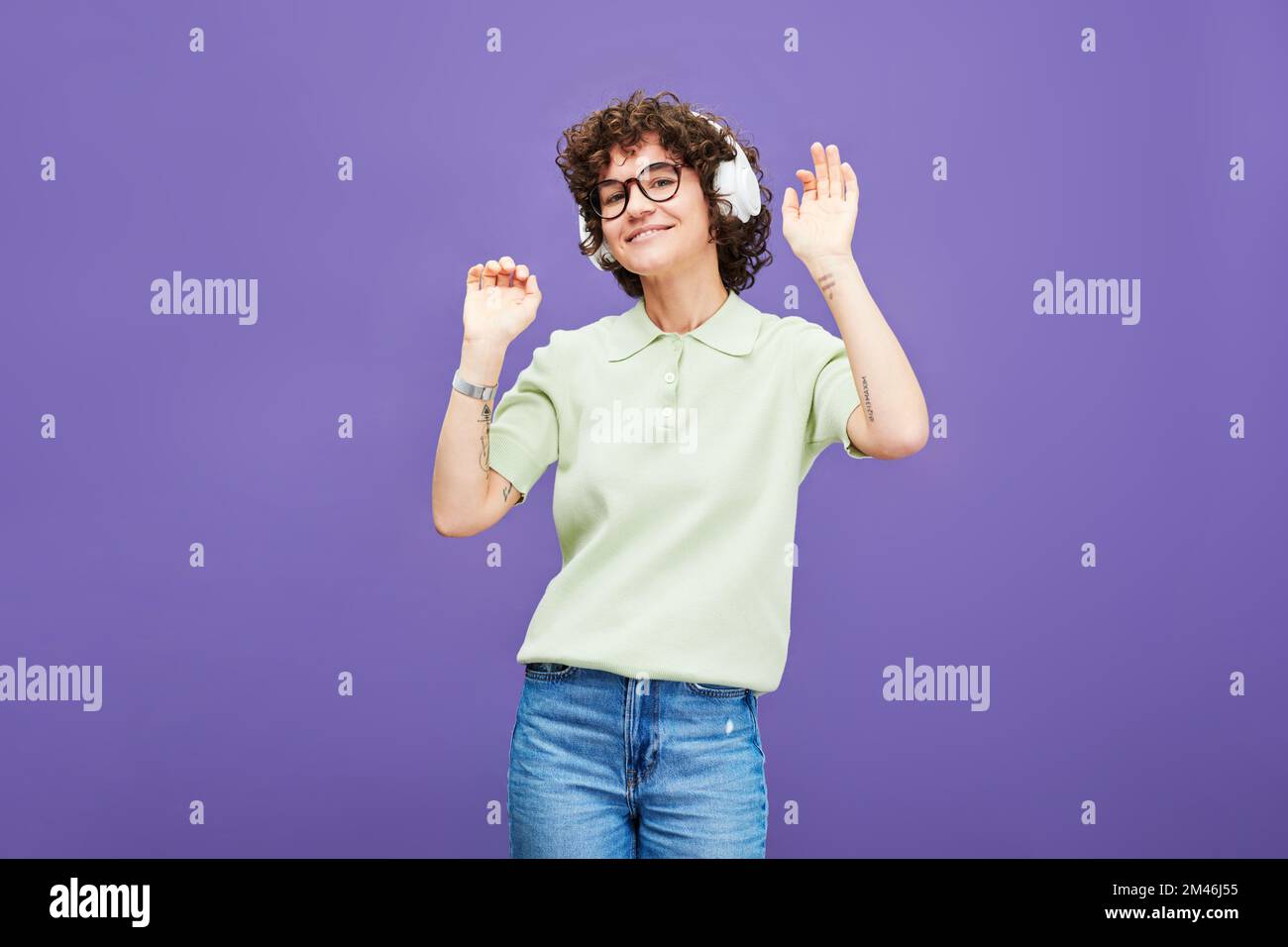 Junge fröhliche Frau in Kopfhörern und Freizeitkleidung, die vor der Kamera auf Lavendel-Hintergrund in Isolation Musik hört und tanzt Stockfoto