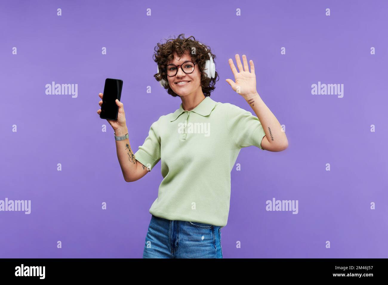Junge fröhliche Frau mit Tattoos auf den Armen, die energiegeladene Musik über Kopfhörer hört und vor der Kamera auf violettem Hintergrund tanzt Stockfoto