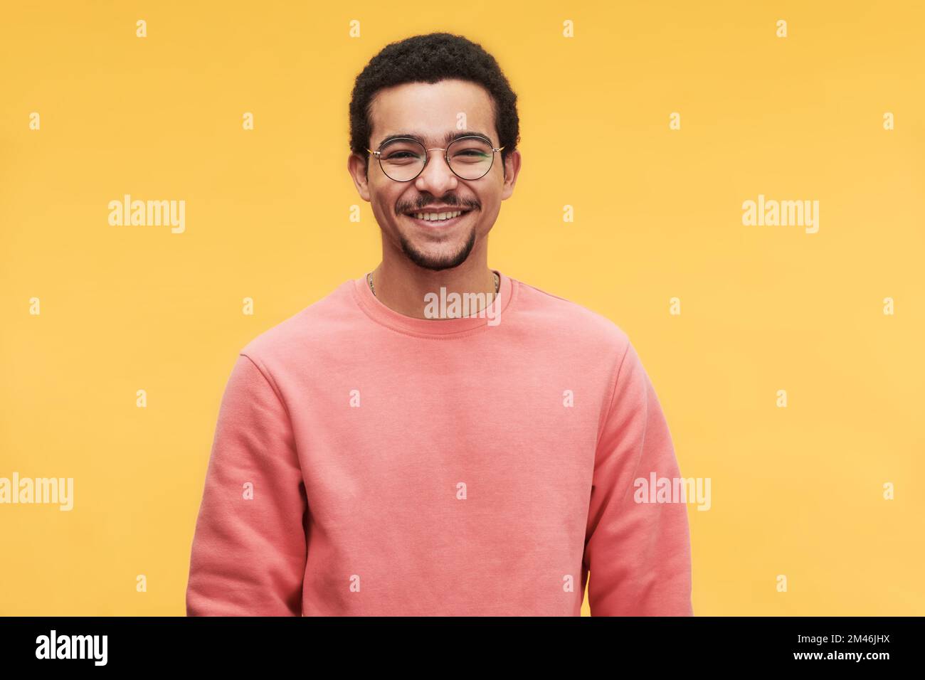 Junger fröhlicher Mann mit Brille und rosa Sweatshirt, der in die Kamera schaut, während er isoliert vor einem lebendigen gelben Hintergrund steht Stockfoto