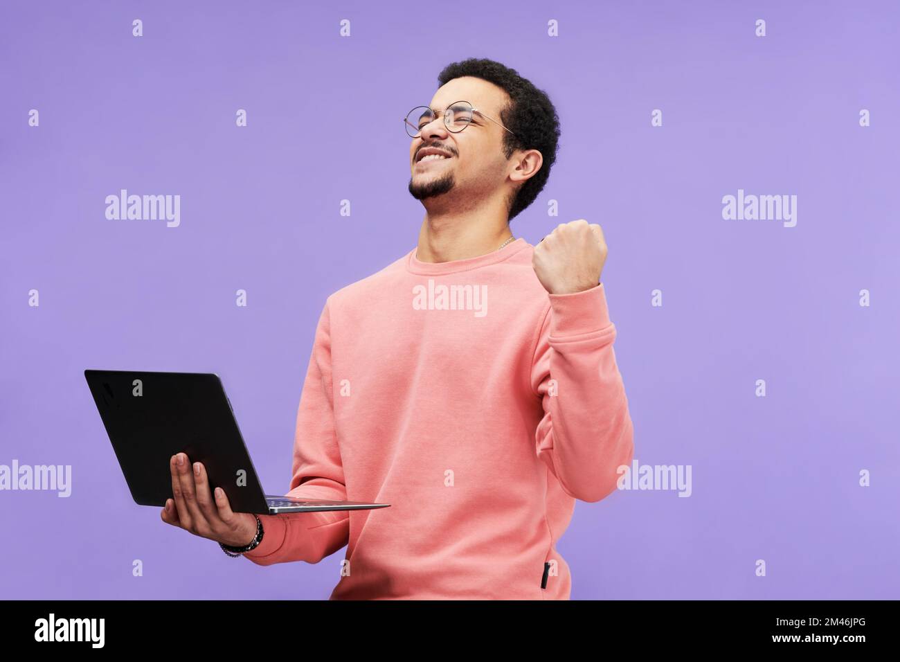 Ekstatischer Typ in pinkfarbener Freizeitkleidung hält die Augen geschlossen, während er den Laptop hält und die Aufregung vor violettem Hintergrund isoliert ausdrückt Stockfoto