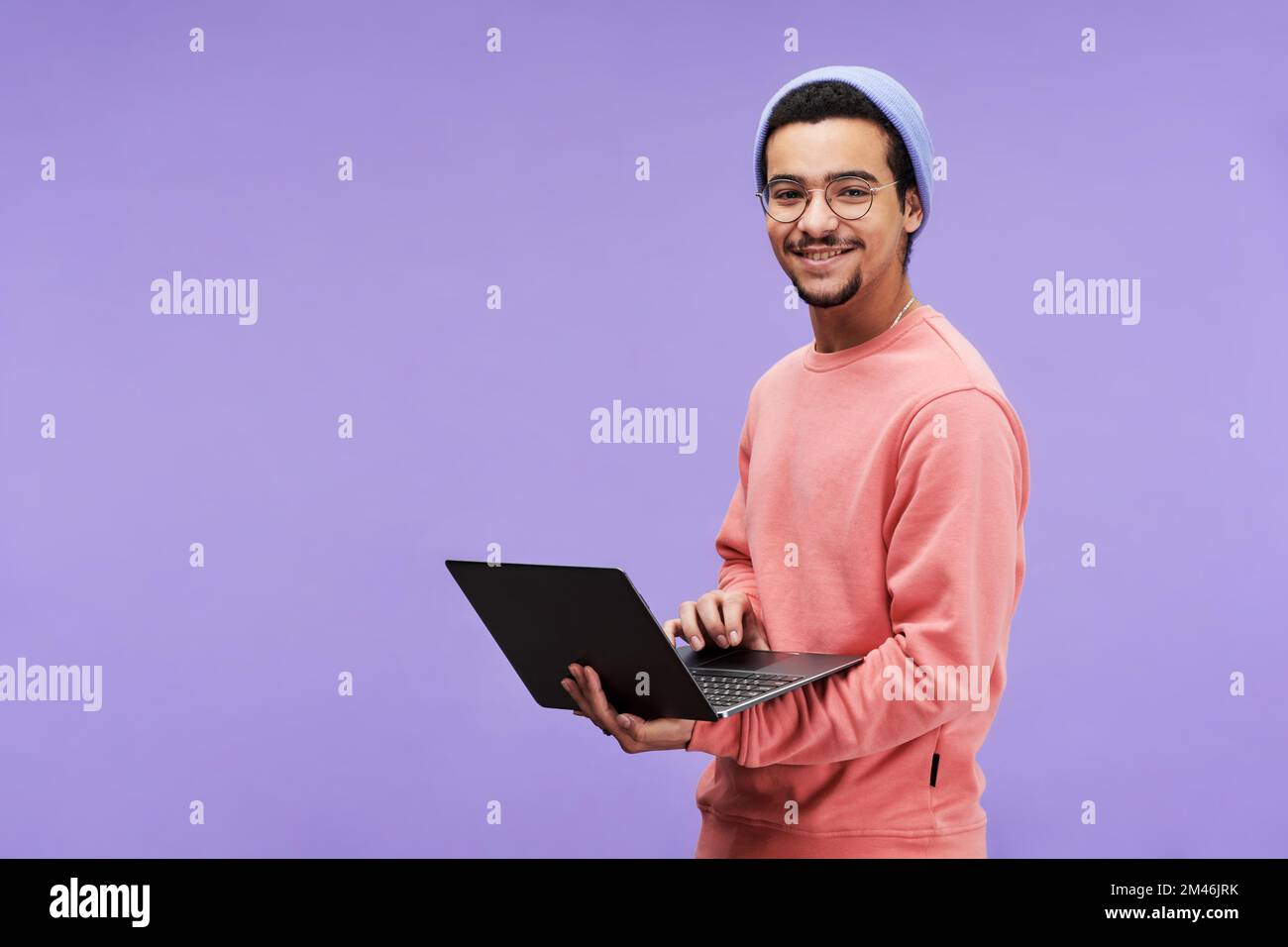 Junger, erfolgreicher Designer oder Freiberufler in Freizeitkleidung, der ein Notebook benutzt und mit einem Lächeln auf die Kamera schaut, während er auf violettem Hintergrund steht Stockfoto