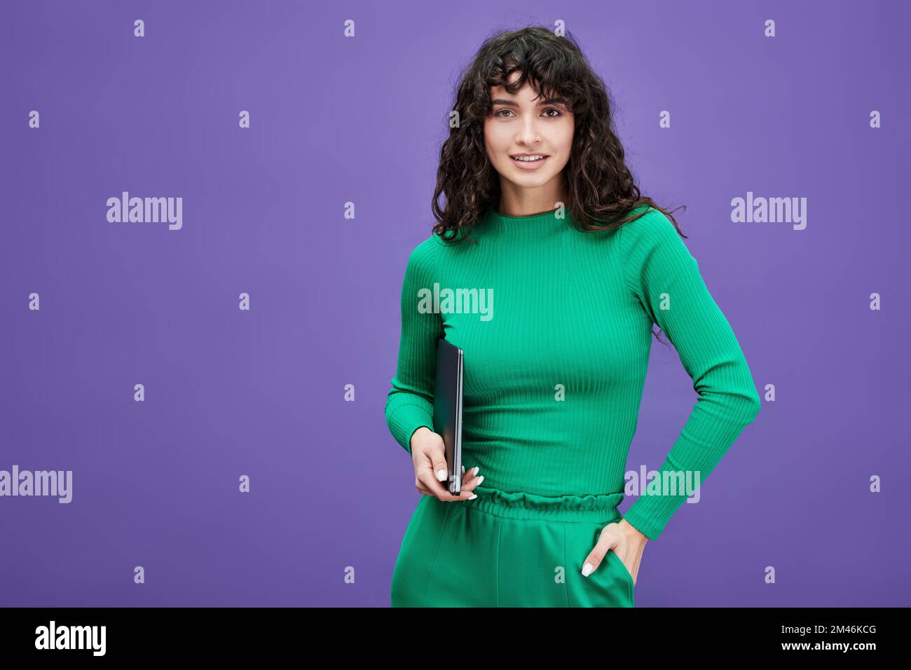 Junge selbstbewusste Mitarbeiterin oder Studentin in grüner Freizeitkleidung, die ihren Laptop hält und auf violettem Hintergrund in die Kamera schaut Stockfoto