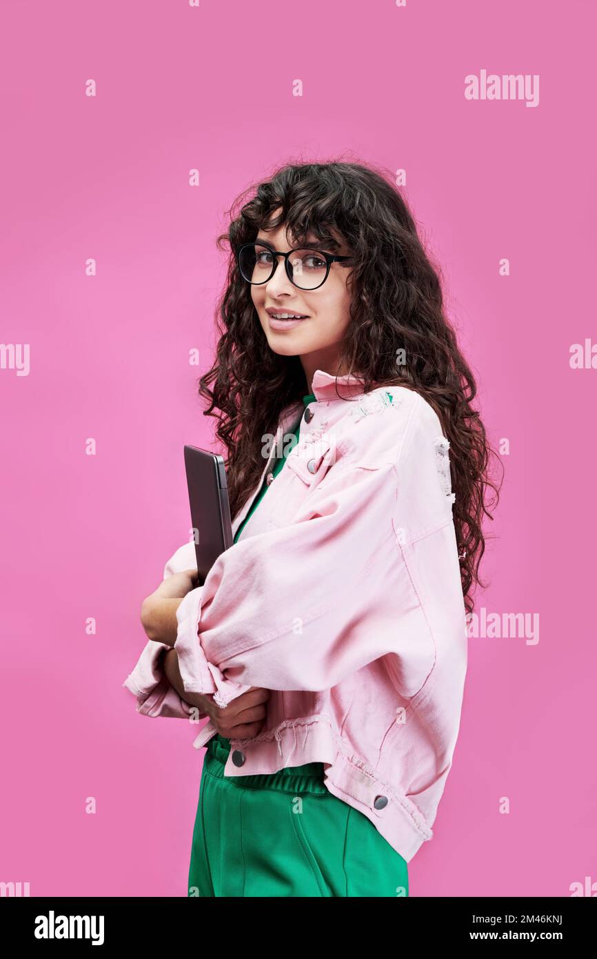 Braune Studentin in Freizeitkleidung mit mobilem Gerät und Blick in die Kamera, während sie während der Fotosession auf pinkfarbenem Hintergrund posiert Stockfoto