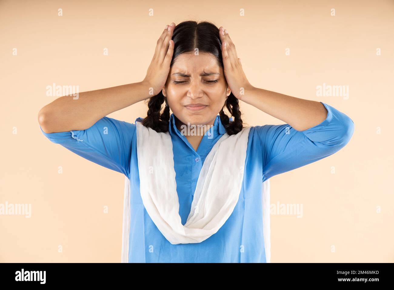 Gestresste und depressive, ländliche indische Schülerin in blauer Schuluniform mit beiden Händen am Kopf isoliert auf beigem Hintergrund, Ne Stockfoto