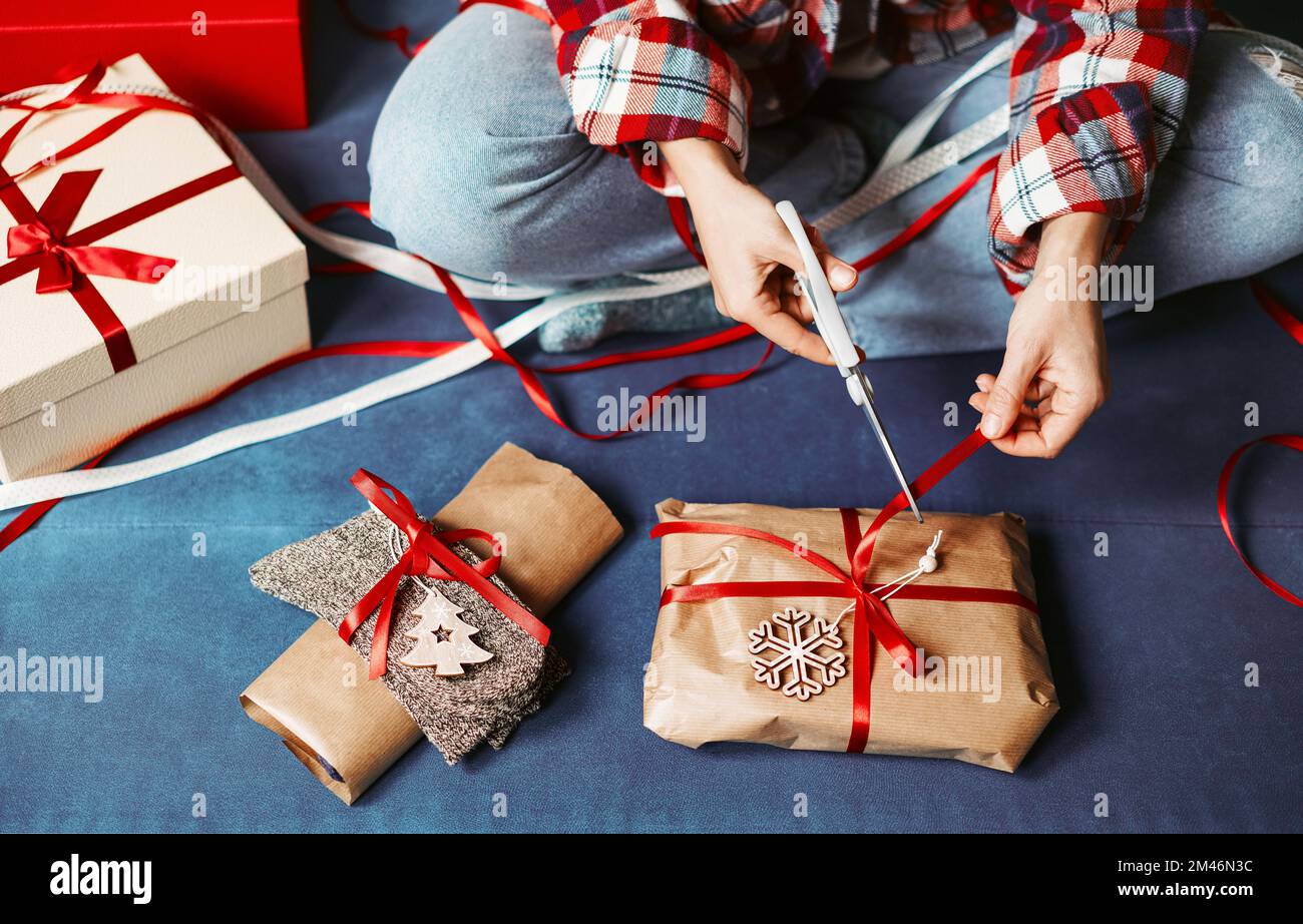 Verpackt Weihnachtsgeschenke mit braunem Handwerkspapier und schneidet das rote Band. Stockfoto