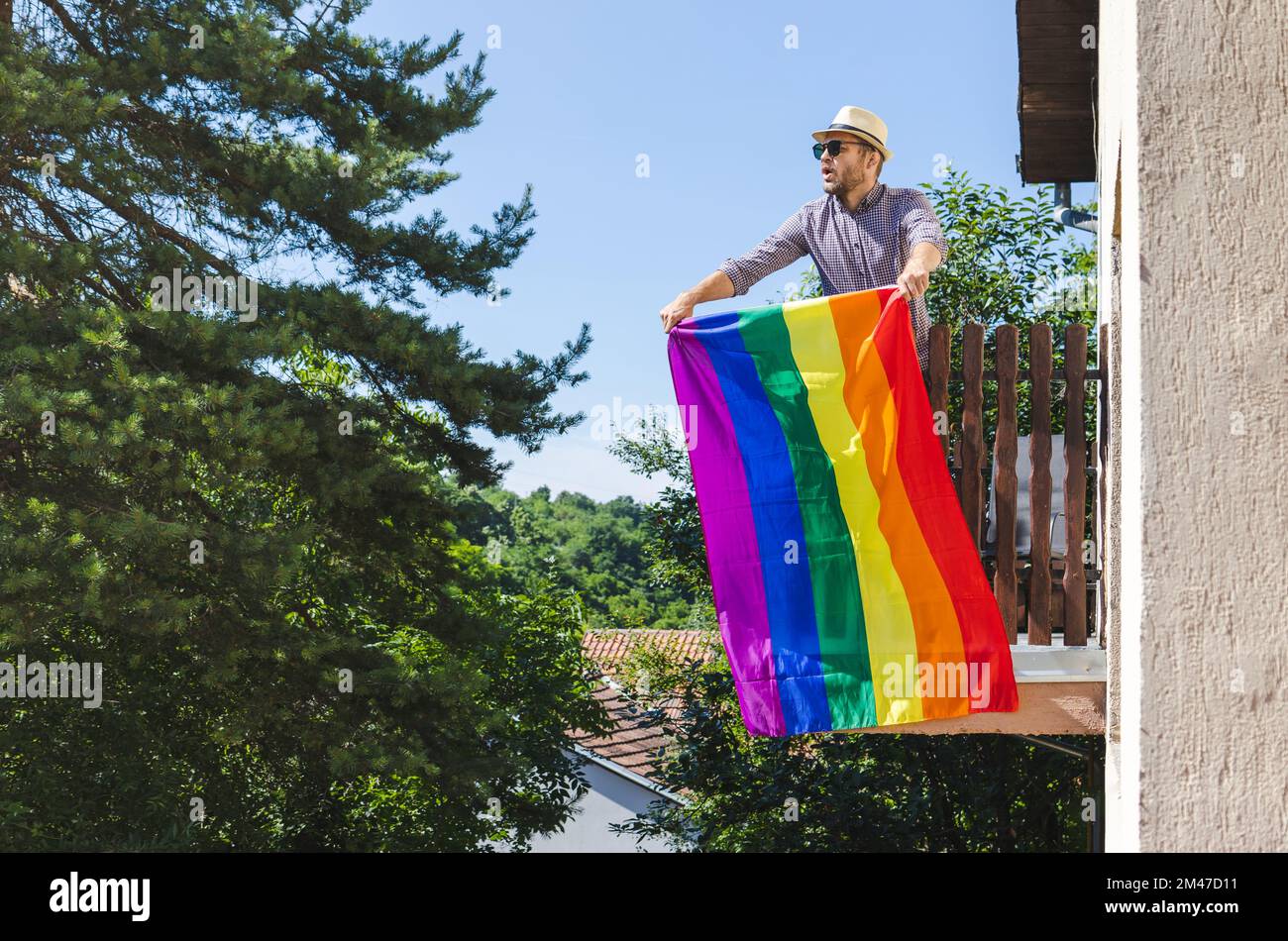 Heterosexueller Mann mit Hut, der auf dem Balkon steht und eine Regenbogenflagge hält und dabei die LGBT-Gemeinschaft, Menschenrechte, Gleichheit und Vielfalt unterstützt Stockfoto