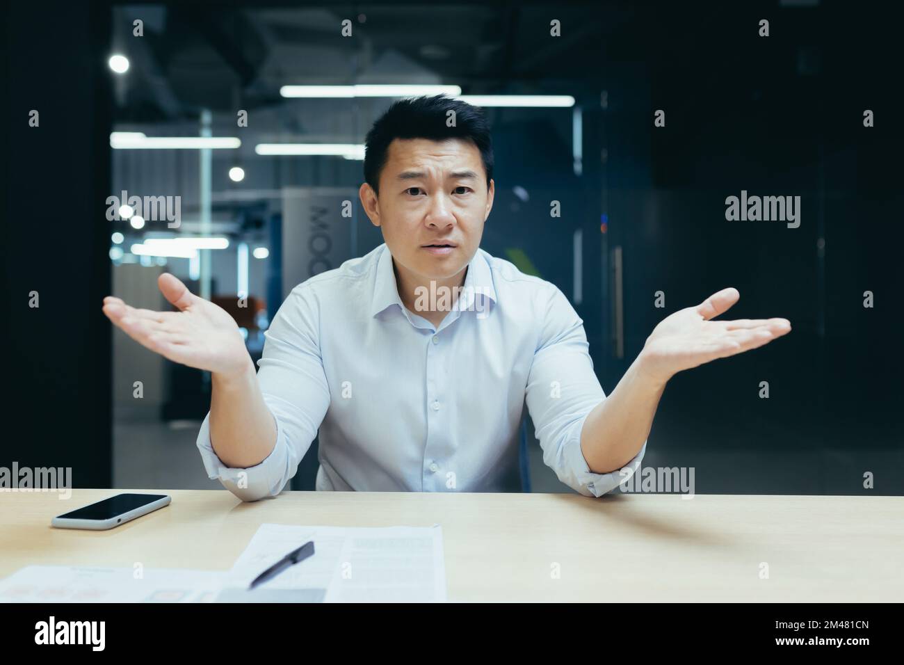 Besorgter und seriöser junger asiatischer Mann, Büroangestellter sitzt am Schreibtisch, spreizt seine Hände, hat Probleme, schaut in die Kamera. Stockfoto