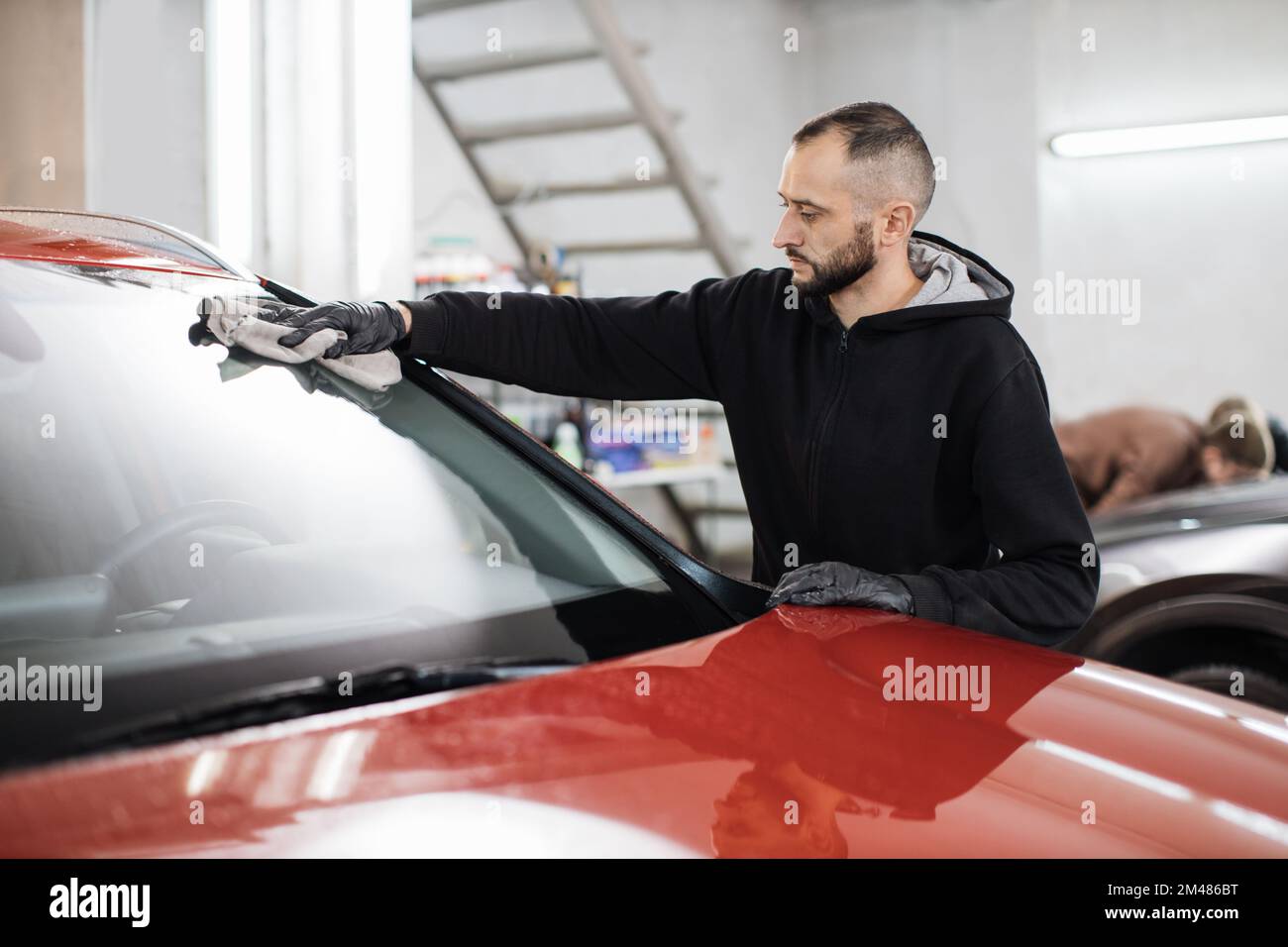 Fahrzeugdetails und Reinigungskonzept. Gut aussehender junger Autowaschanlagenarbeiter, trägt schwarze Handschuhe, reinigt und poliert Autofenster mit Mikrofasertuch. Stockfoto