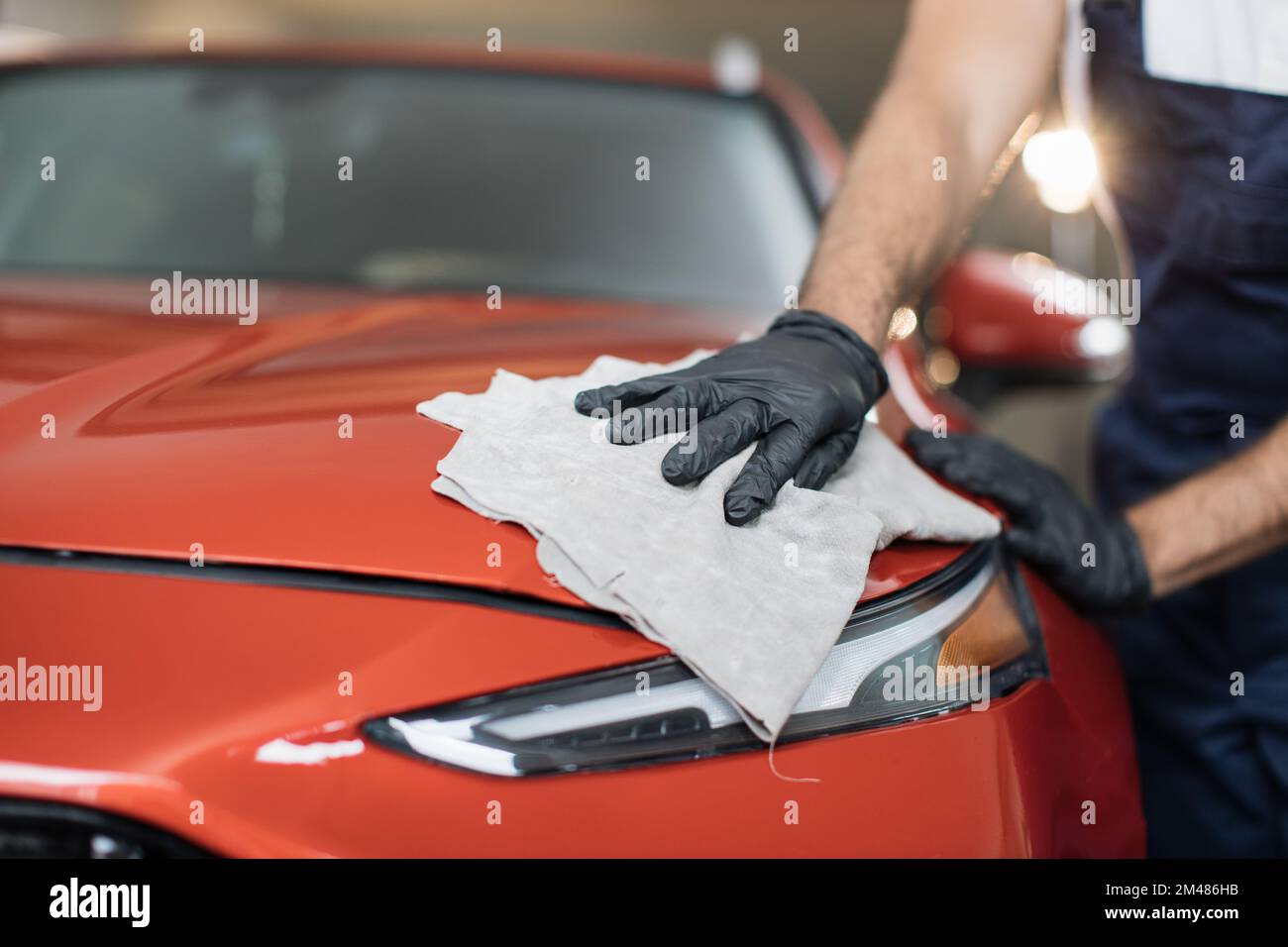 Zugeschnittenes Bild der Hand eines männlichen professionellen Autowaschanlagenarbeiters in schwarzen Gummihandschuhen, der die graue Mikrofaser hält und die Motorhaube und den Scheinwerfer eines luxuriösen roten Autos poliert. Stockfoto