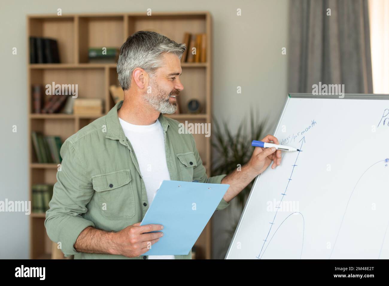 Lächelnder älterer, weißer Mann Lehrer schreibt an Tafel und nimmt Videolehrgang im Wohnzimmer auf Stockfoto