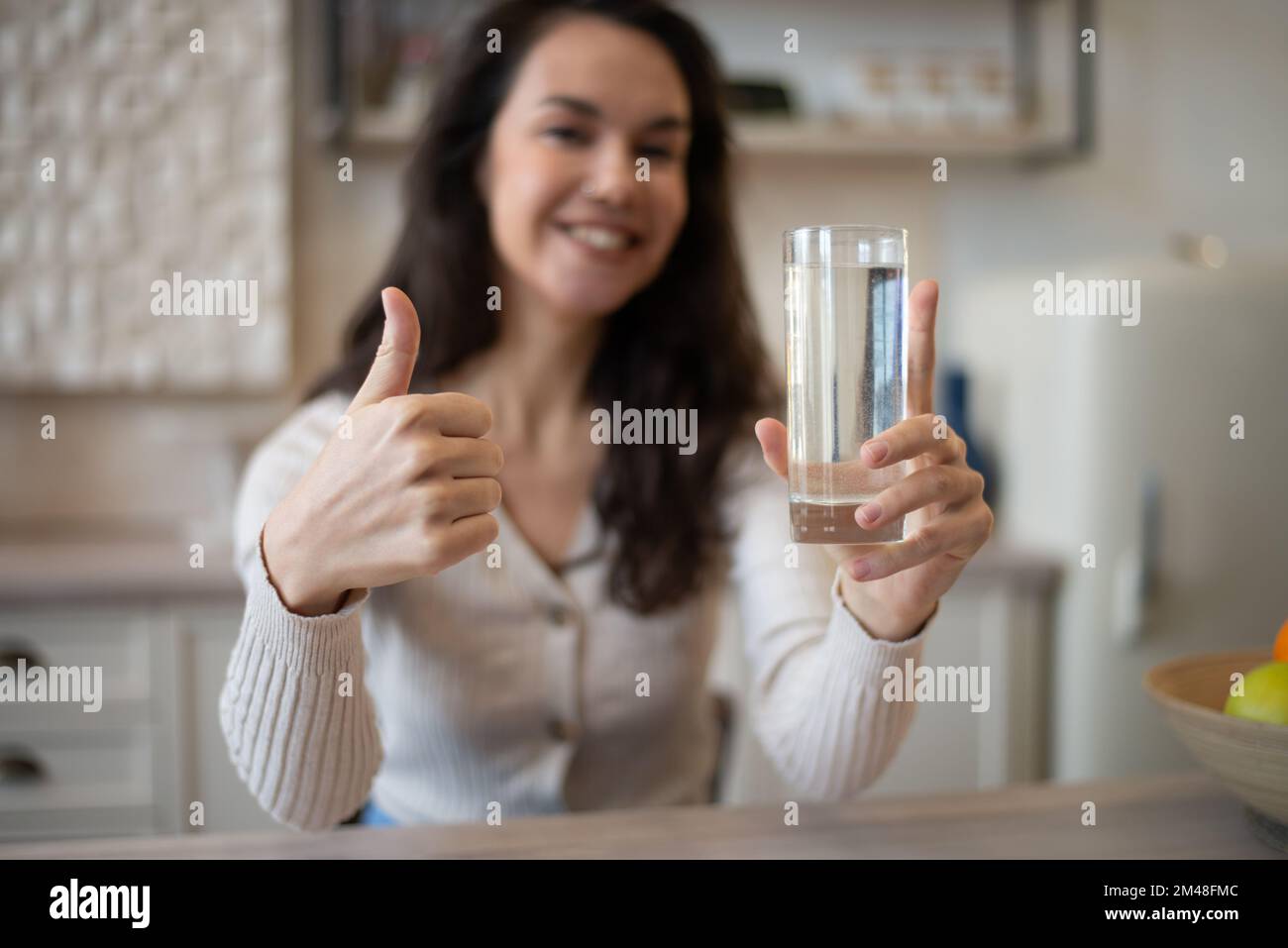 Aufgeregte kaukasische Dame hält ein Glas Wasser und zeigt Daumen-hoch-Geste, sitzt am Tisch in der Küche, selektive Fokussierung Stockfoto