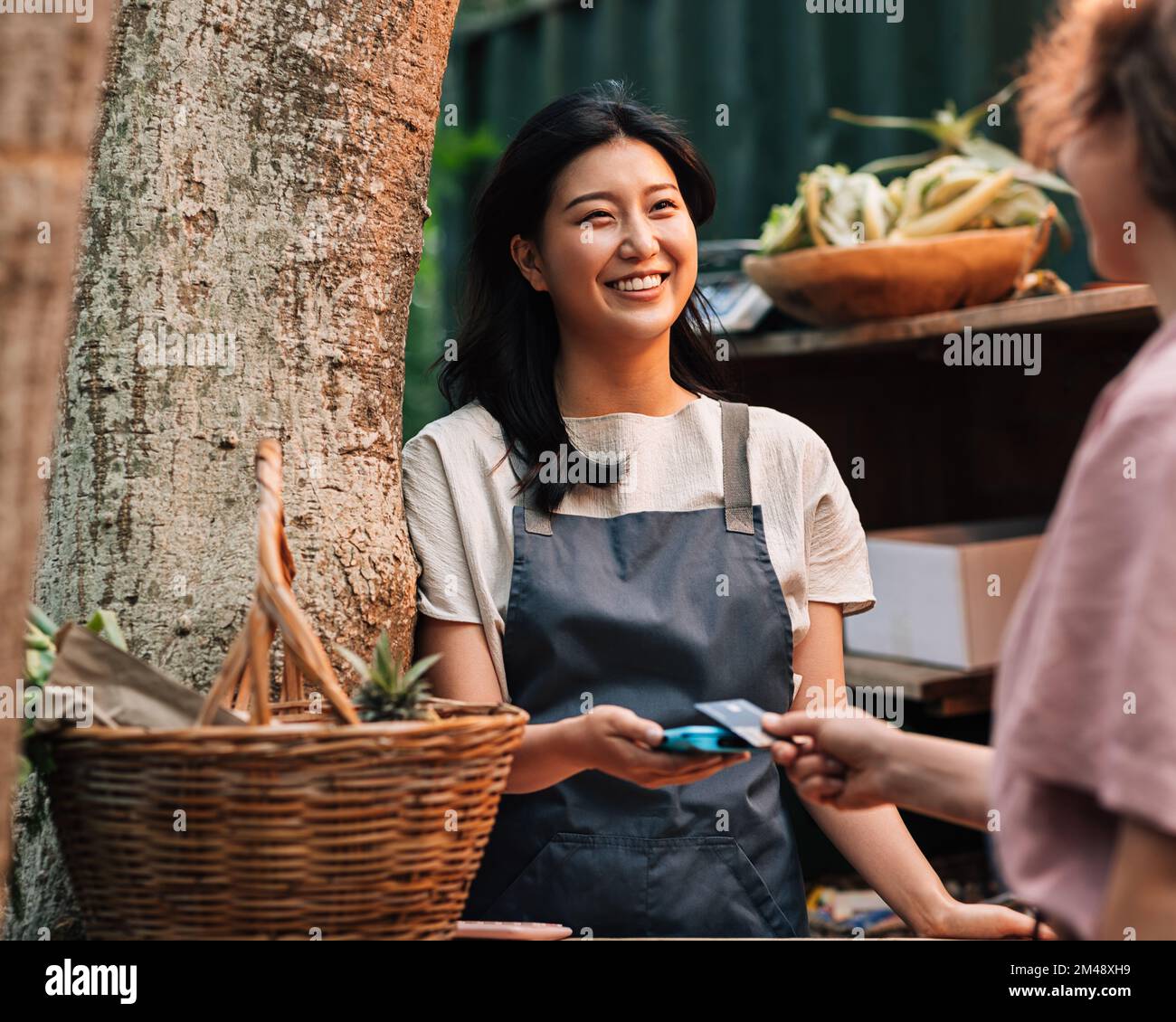 Glückliche Asiatin in einer Schürze, die auf dem Markt bezahlt wird. Anbieter mit einem Kartenautomaten, der den Kunden lächelt und ansieht. Stockfoto