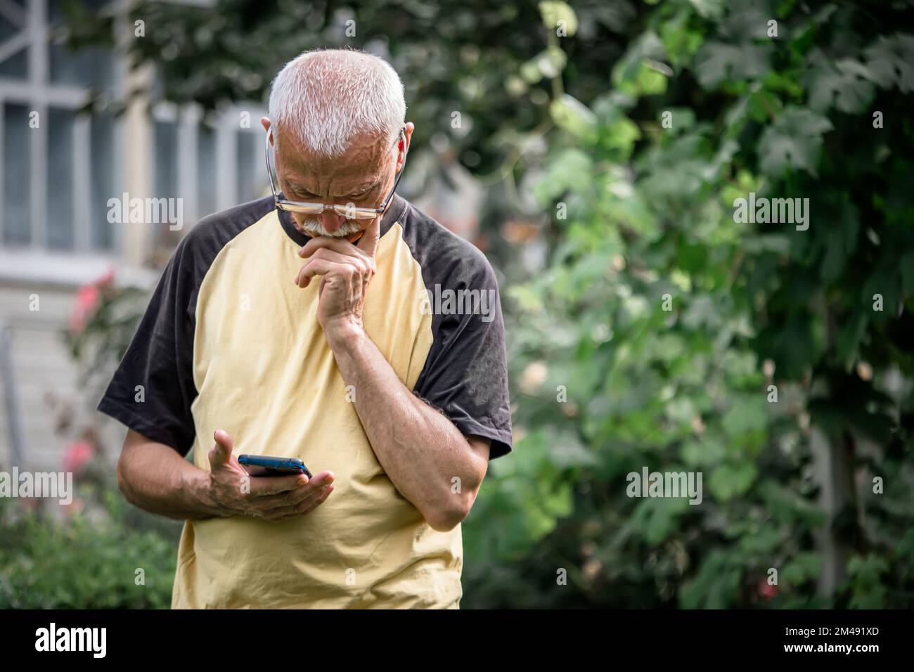 Ein grüblerischer alter Bauer vor einem alten Haus nimmt aus der Ferne an einem Smartphone-Webinar über alternative Therapie der psychischen Gesundheit Teil Stockfoto