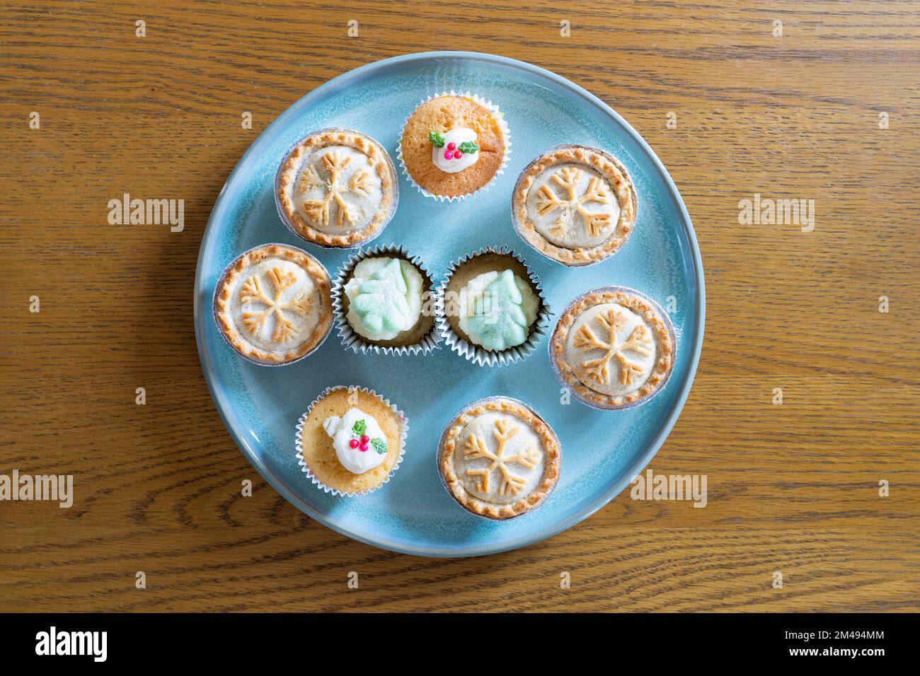 Hackkuchen mit Schneeflockenmuster und Tassentörtchen mit essbaren Stachelblättern und Beeren und Weihnachtsbäumen auf einem blauen Teller auf einem Eichenholztisch Stockfoto