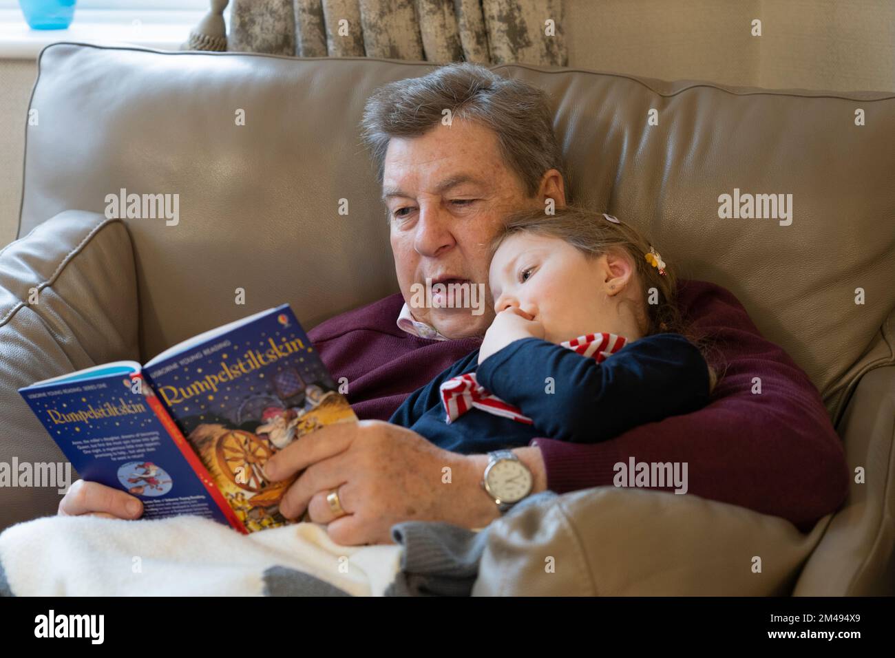 Ein Großvater, der seiner vierjährigen Enkelin, die aufmerksam zuhört, das klassische deutsche Märchenbuch der Kinder Rumpelstilzchen vorliest. UK Stockfoto