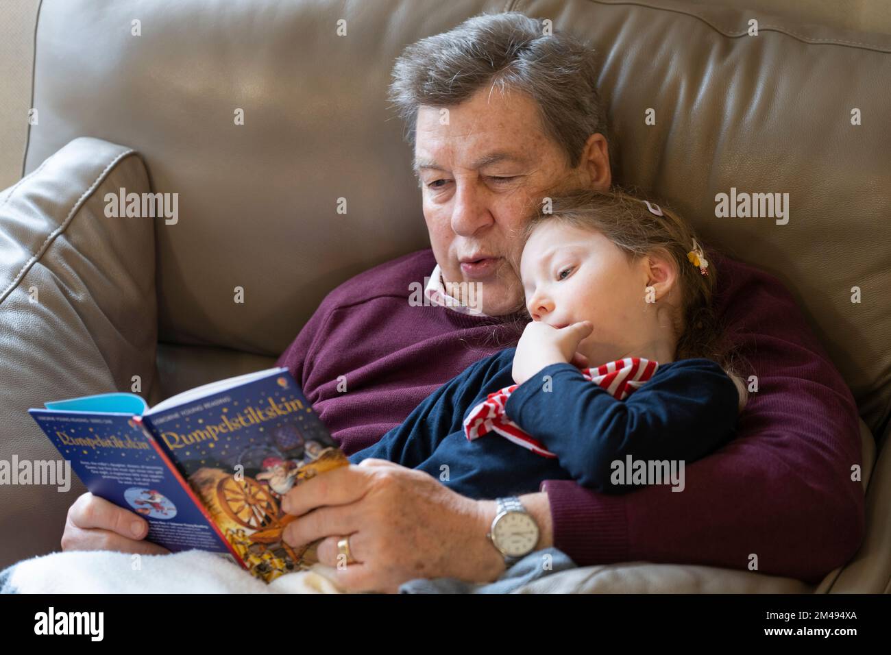 Ein Großvater, der seiner vierjährigen Enkelin, die aufmerksam zuhört, das klassische deutsche Märchenbuch der Kinder Rumpelstilzchen vorliest. UK Stockfoto