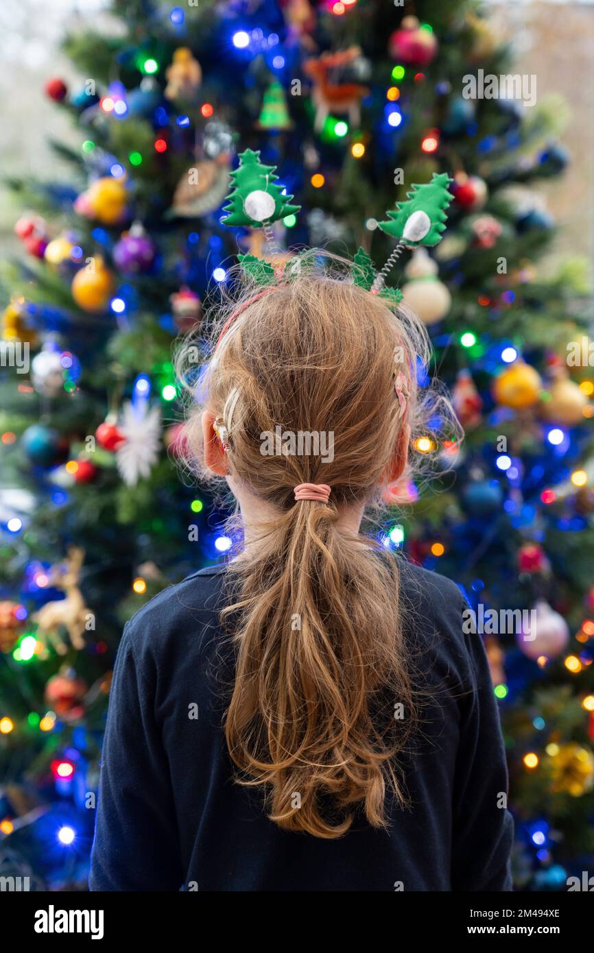 Ein kleines Mädchen (4), das auf einen Weihnachtsbaum schaut, der mit Feenlichtern und Kugeln dekoriert ist. Thema: Weihnachten, Weihnachtsgrüße, Kinderwunder Stockfoto