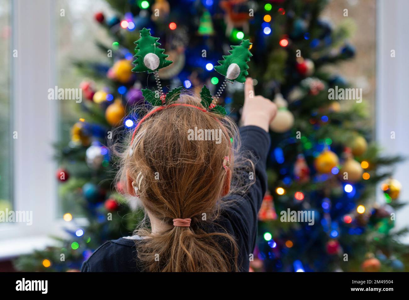 Ein kleines Mädchen (4), das auf einen Weihnachtsbaum zeigt, der mit Lichtern und Kugeln dekoriert ist. Thema: Weihnachten, Weihnachtsgrüße, Kinderwunder Stockfoto