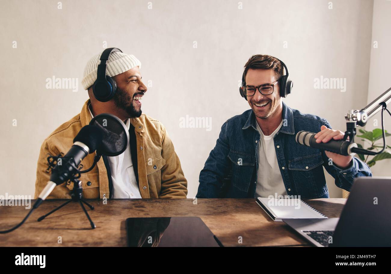 der college-Radio-dj lacht und hat eine gute Zeit auf Sendung. Zwei glückliche junge Männer, die eine Live-Audioübertragung in einem Studio aufnehmen. Fröhliche Inhaltsersteller Stockfoto