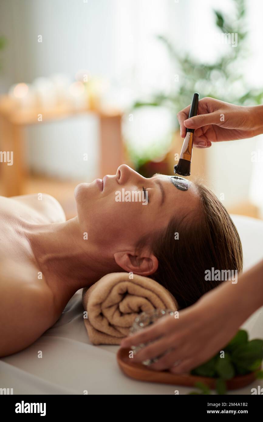 Asthetikerin, die Tonmaske auf das Gesicht einer erwachsenen Klientin aufträgt Stockfoto