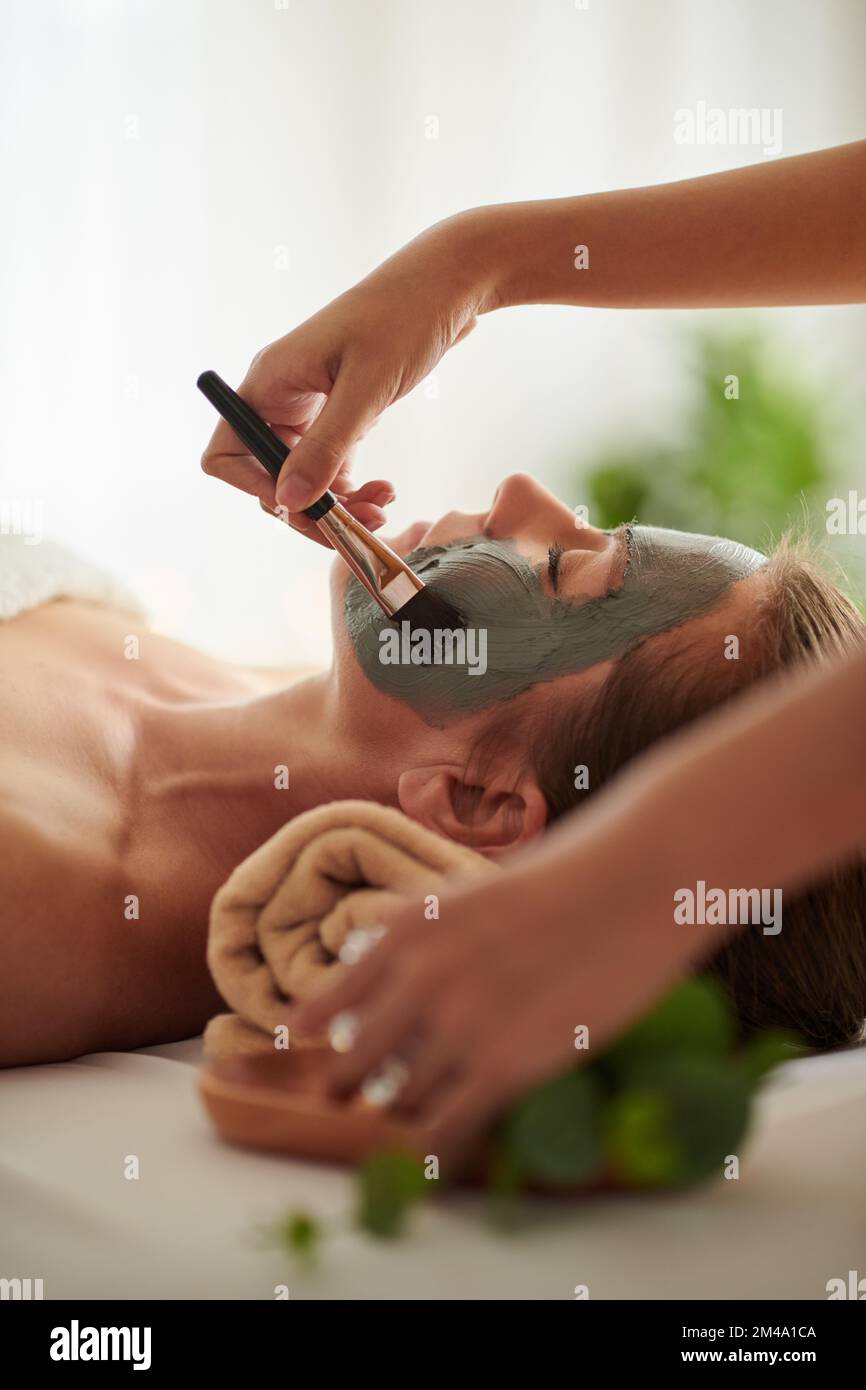 Kosmetikerin trägt schwarze Tonmaske auf das Gesicht des Kunden im Schönheitssalon Stockfoto
