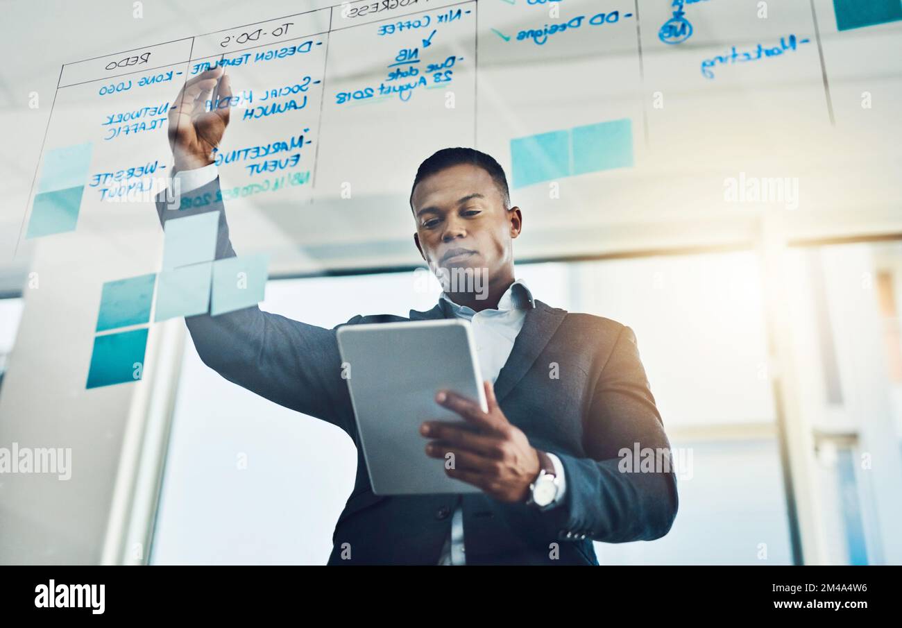 Technologie hilft ihm, seine Arbeit schneller zu erledigen. Ein junger Geschäftsmann, der ein digitales Tablet benutzt, während er sich Notizen an eine Glaswand im Büro schreibt. Stockfoto