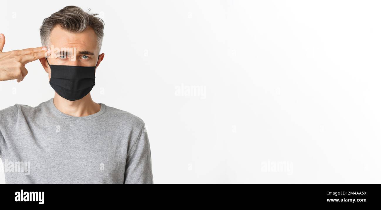 Konzept von covid-19, sozialer Distanzierung und Quarantäne. Nahaufnahme eines traurigen Mannes mittleren Alters in schwarzer medizinischer Maske, mit der Fingerpistole auf den Kopf zeigend und schauend Stockfoto