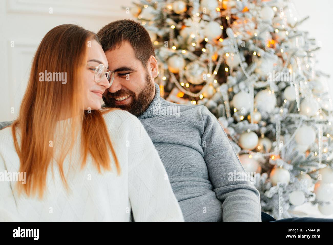 Ein verliebtes Paar umarmt und küsst sich in der Nähe des Weihnachtsbaums im dekorierten Inneren des Weihnachtsbaums zu Hause an einem Weihnachtsurlaub im Winter. Ein Mann Stockfoto