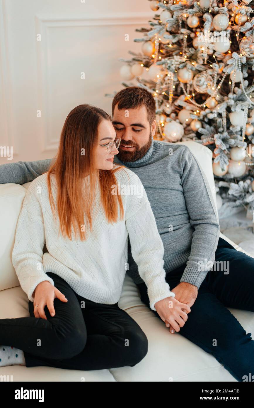 Ein verliebtes Paar umarmt und küsst sich in der Nähe des Weihnachtsbaums im dekorierten Inneren des Weihnachtsbaums zu Hause an einem Weihnachtsurlaub im Winter. Ein Mann Stockfoto