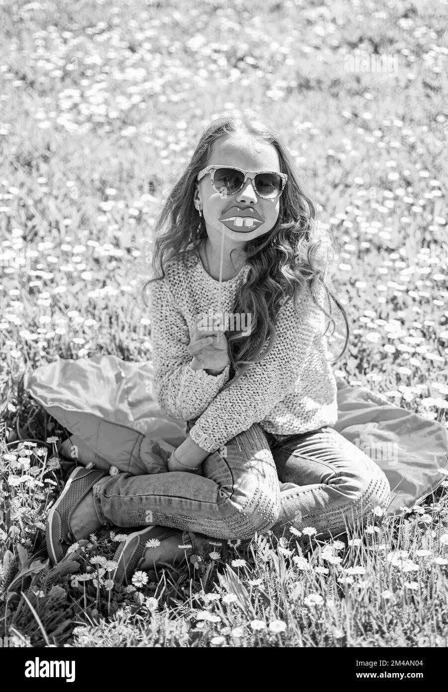 Das Mädchen sitzt auf Gras am Grassplot, grüner Hintergrund. Kinder, die mit Brillen posieren, lächelnde Lippen aus Pappe für eine Fotosession auf der Wiese. Humor-Konzept Stockfoto