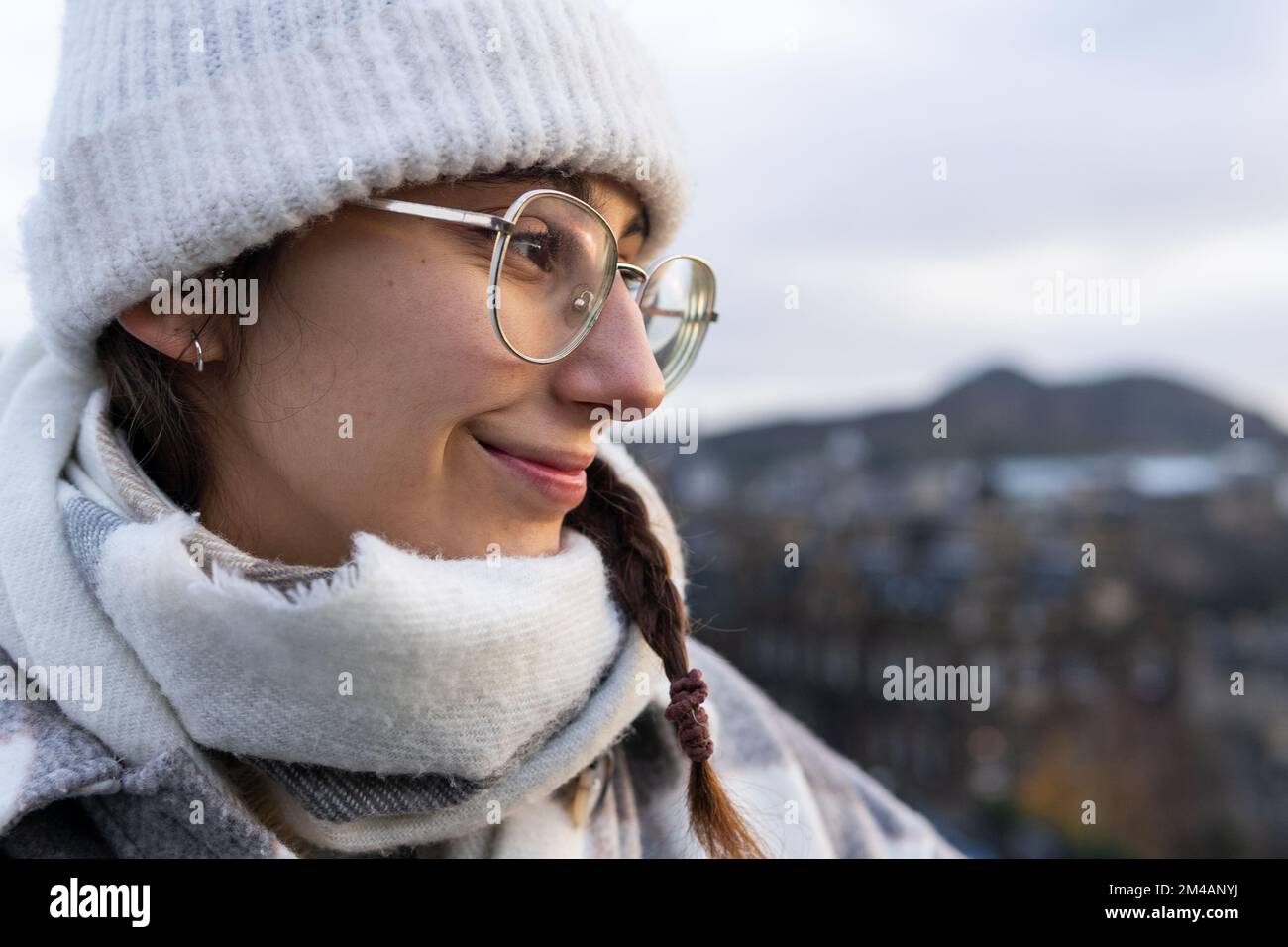 Positive junge Frau mit warmem Hut und Schal mit einer lächelnden Brille und Blick auf den verschwommenen Hintergrund von Edinburgh an einem grauen Tag in Schottland Stockfoto
