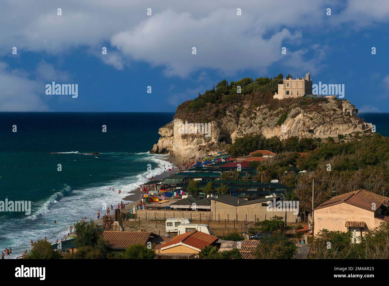 Urlauber paddeln im Tyrrhenischen Meer am Sandstrand unter dem Heiligtum Santa Maria dell’Isola, das nach Erdbeben wieder aufgebaut wurde und sich auf einem felsigen Vorsprung unterhalb der Klippenstadt Tropea auf den westlichen Kosten von Kalabrien in Süditalien befindet. Der Felsen, der einst eine Insel war, ist seit vielen Jahrhunderten eine Klosterstätte. Stockfoto