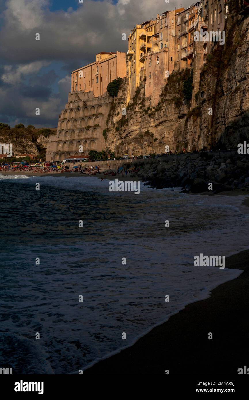 Am späten Abend im August Sonnenlicht und Schatten, während das dunkle Tyrrhenische Meer einen Strand umrundet, der immer noch mit Urlaubern beschäftigt ist, unter der spektakulären Stadt und dem Ferienort Tropea auf den Klippen an der Costa degli Dei (Küste der Götter) in Westkalabrien, Süditalien. Stockfoto