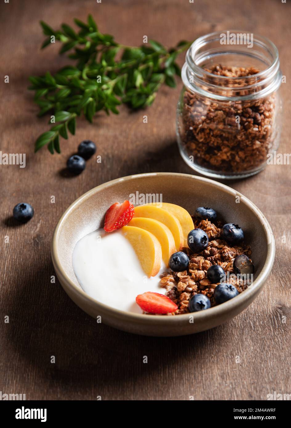 Frischer Naturjoghurt mit hausgemachtem Müsli, Pfirsich, Erdbeere und Heidelbeere in einer Schüssel auf dunklem Holzhintergrund. Das Konzept des gesunden an Stockfoto