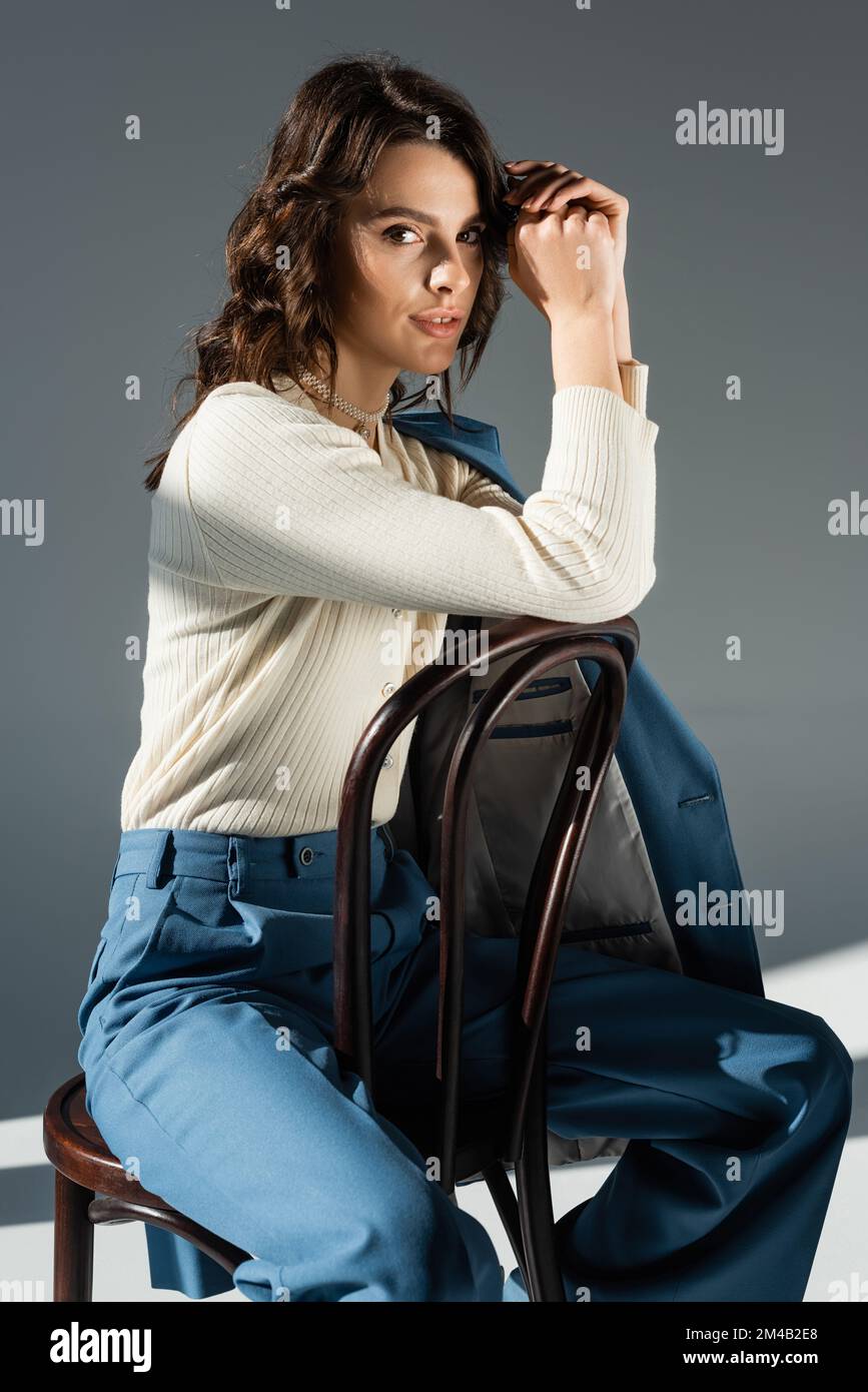 Positive Frau in weißem Pullover und blauer Hose, die auf einem Stuhl sitzt und auf grauen Hintergrund in die Kamera schaut Stockfoto