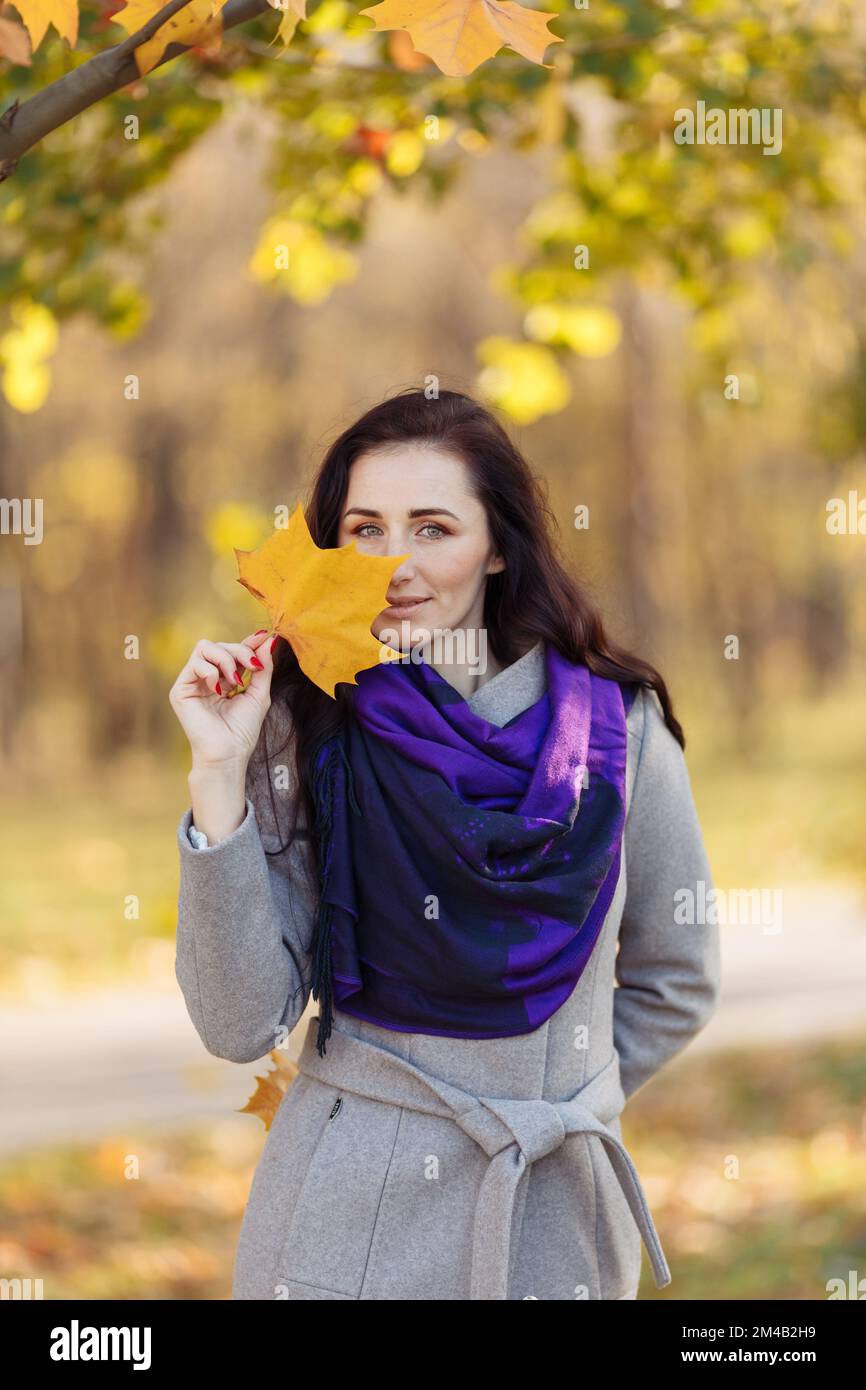 Ein hübsches Mädchen mit dunklen Haaren, einem nassen Mantel und einem lila Schal hält einen Brief. Unscharfer Hintergrund und unscharfe Blätter sind oben sichtbar Stockfoto